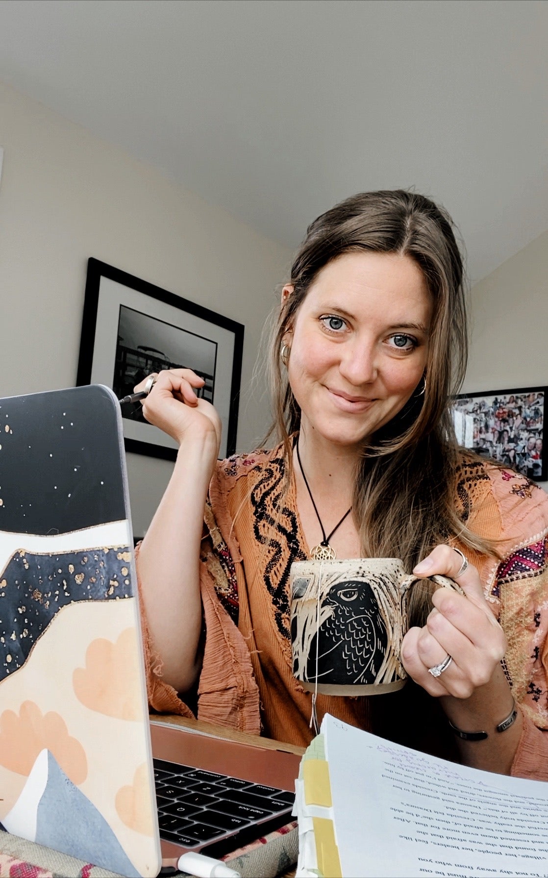 Author and screenwriter, Emily Prescott, before her open laptop in a burnt orange boho top holding a tea mug carved with a hawk.