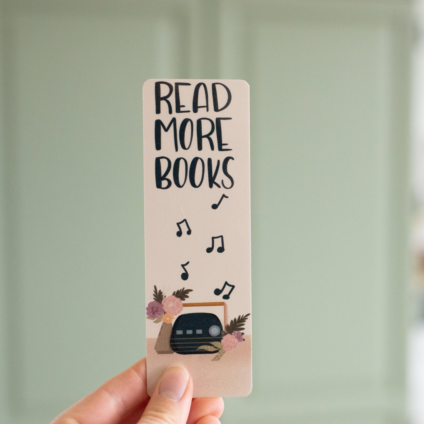 Beige bookmark featuring a navy blue vintage radio beside a vase of boho florals. Music notes rise to meet the block text "Read More Books"