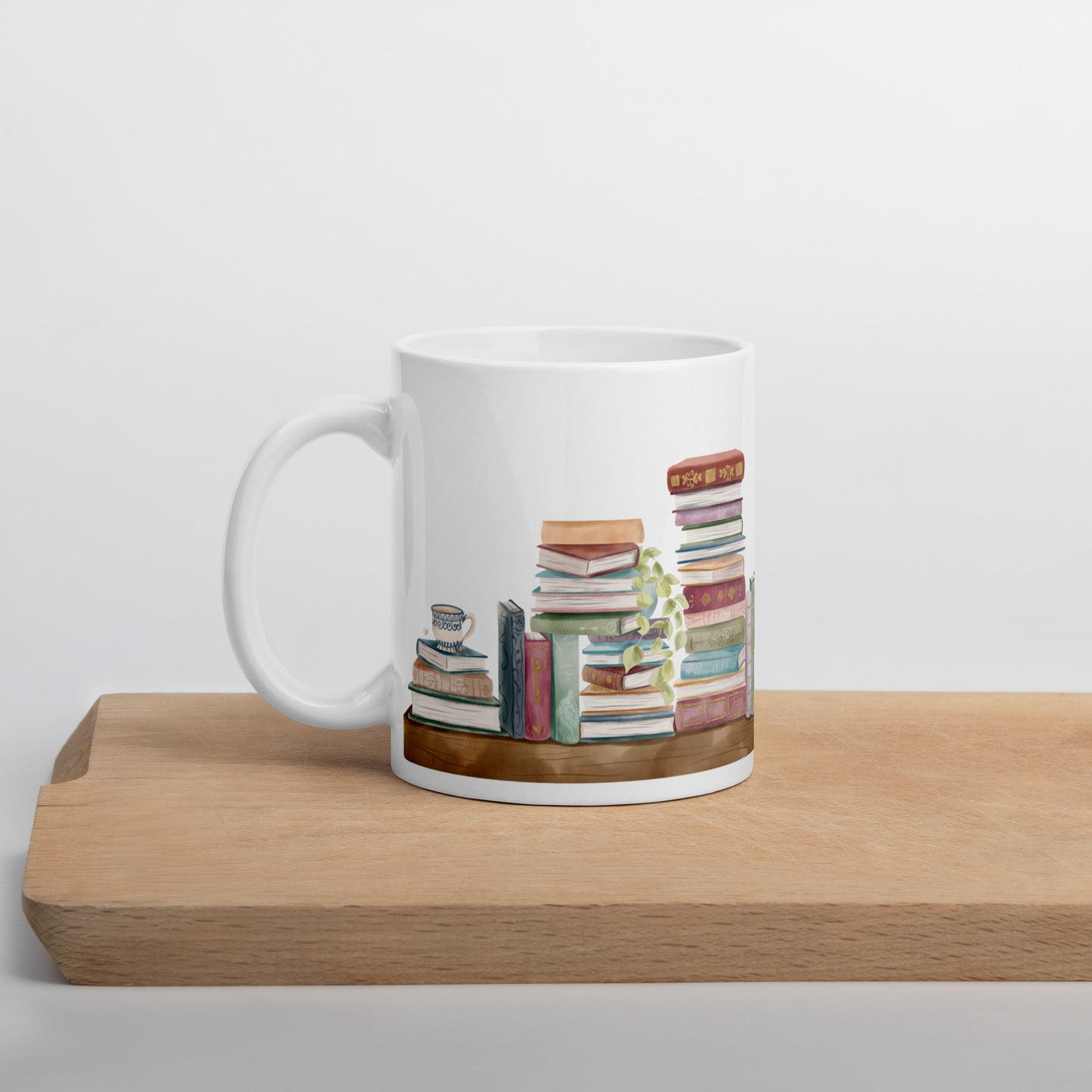 A white ceramic mug for book lovers featuring a wrap around design with stacks of watercolor illustrated books along a shelf.