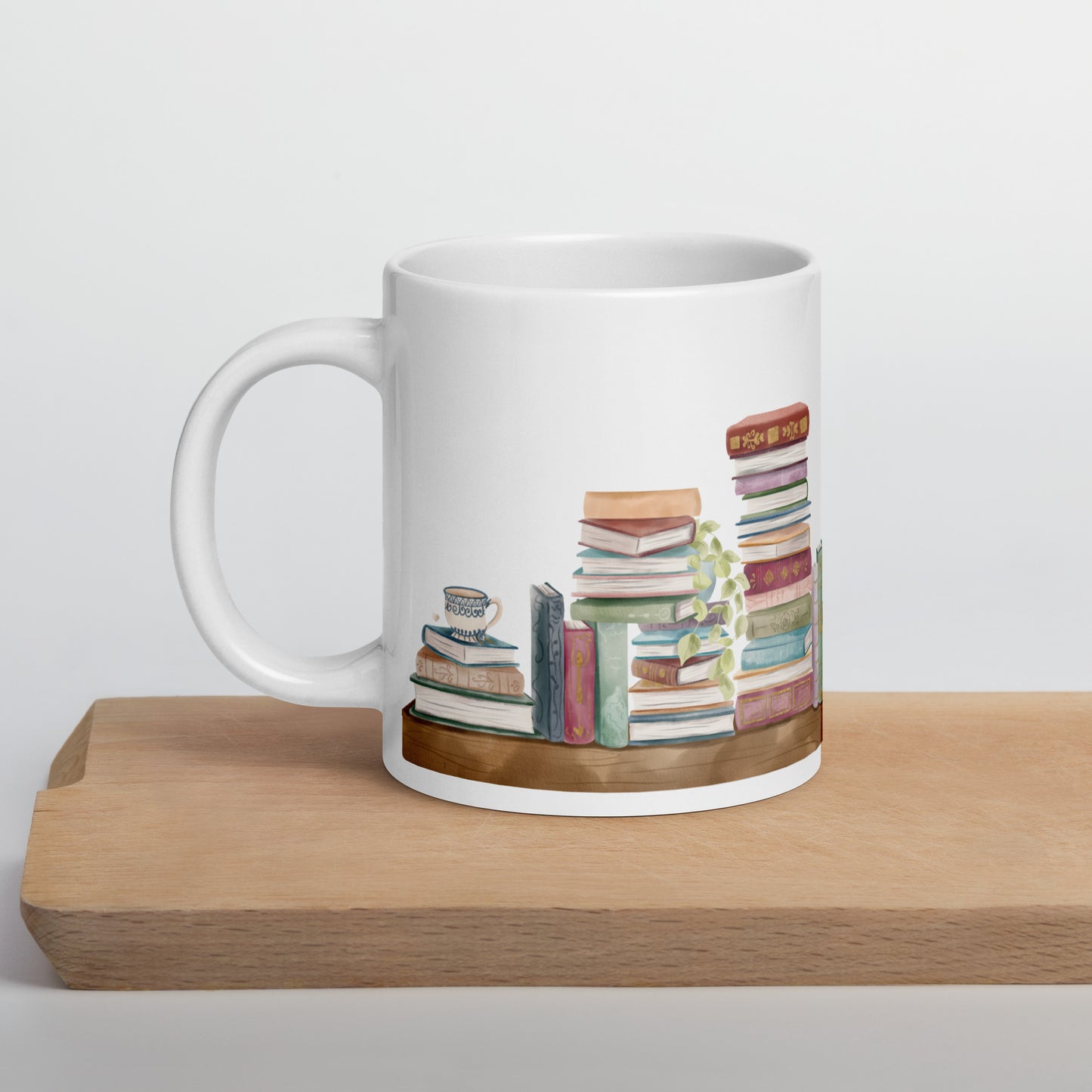 A white ceramic mug for book lovers featuring a wrap around design with stacks of watercolor illustrated books along a shelf.